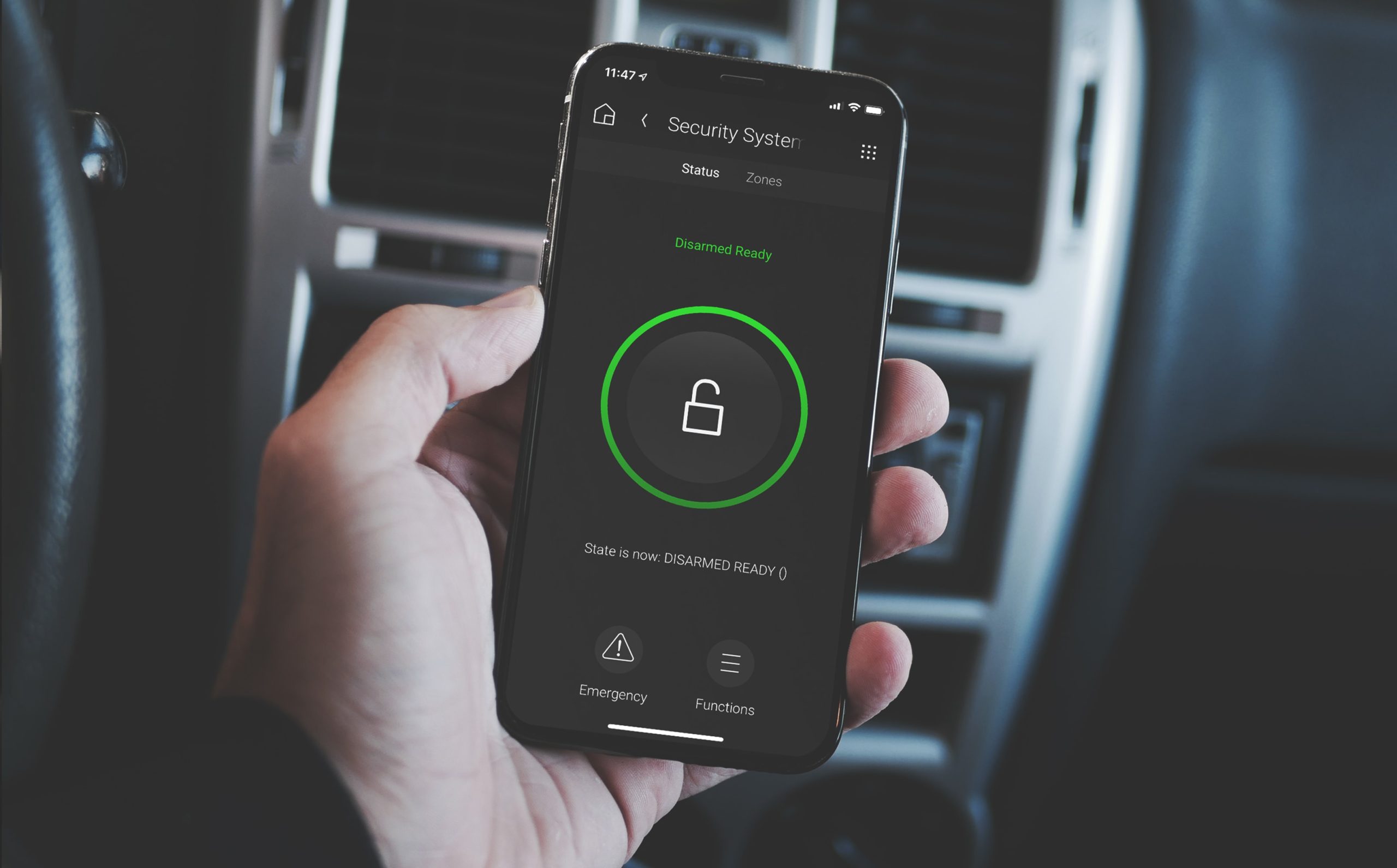 A imagem mostra a mão de uma pessoa segurando um celular. Na tela, é possível visualizar as opções concedidas por um aplicativo de alarme residencial. Ao fundo, o painel de um carro.