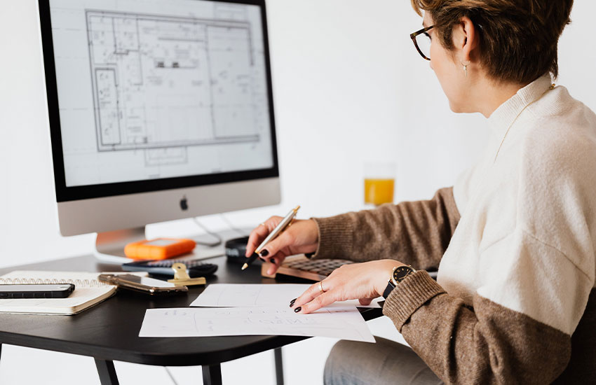 A imagem mostra uma mulher sentada em frente a uma mesa com computador. Ela está de costas, tem cabelos curtos, usa óculos e veste uma blusa de manga longa. A mulher olha para a tela do computador, na qual está a planta de uma casa, e faz desenhos em um papel à sua frente.