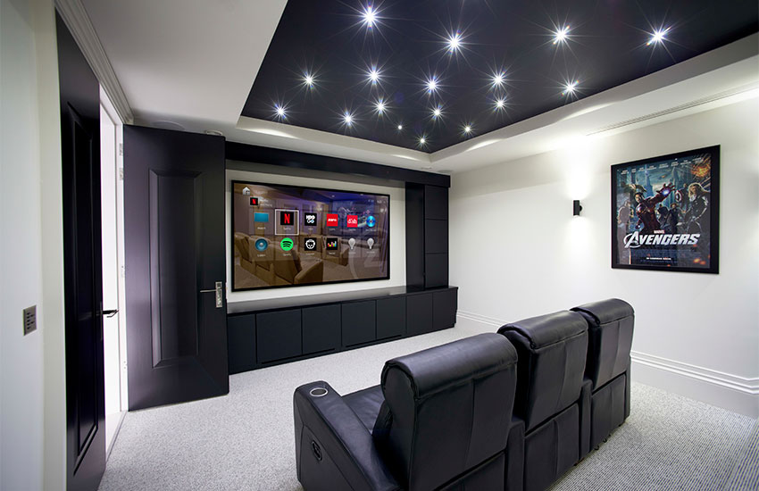 A imagem mostra um ambiente de sala de cinema. Ele tem duas poltronas pretas, uma estante preta e, ao centro do móvel, uma tela grande com ícones de serviços de streaming. No teto, luzes estão brilhando. Na parede à direita há um poster do filme Avengers. À esquerda está uma porta aberta.