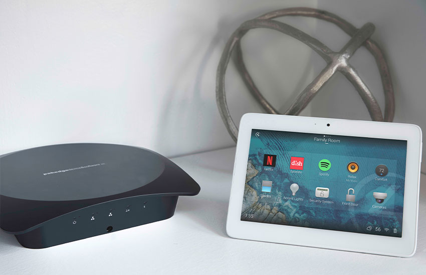 A imagem mostra um roteador de internet preto, ao lado de um tablet branco. Ambos estão posicionados sobre uma superfície branca. Ao fundo está um objeto de decoração esférico e metálico.