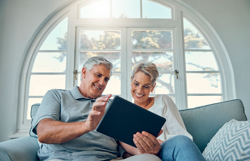 Na imagem, há dois idosos segurando um tablet preto. Eles estão sentados em um sofá e há uma grande janela atrás deles.