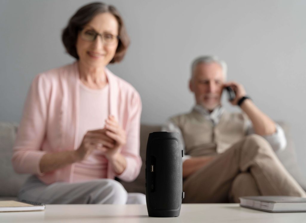 A imagem mostra dois idosos sentados em um sofá. Um deles fala ao telefone e outro sorri enquanto olha para uma caixa de som que está sobre uma mesa de centro.